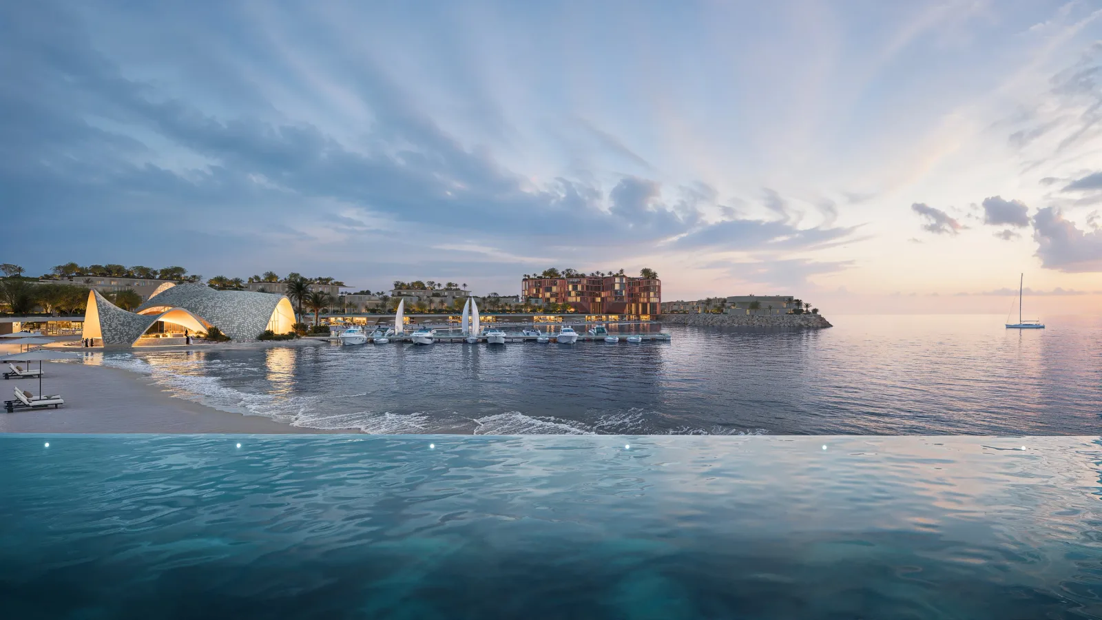 An infinity pool above the beach at dusk, looking across to the lit pavilion and the boat jetty